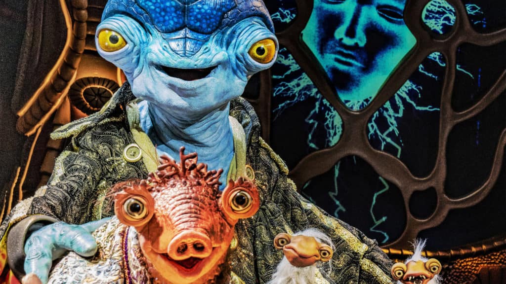 Ned, the blue alien host from Disney+’s "Earth to Ned," dressed in his signature ornate coat and sporting striking yellow eyes, stands center stage against a dazzling cosmic backdrop, cradling two smaller, vibrant alien companions—capturing the whimsical energy of a live Disney Parks meet-and-greet moment.