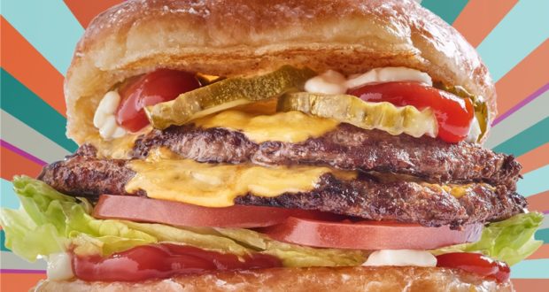 A stacked double cheeseburger loaded with lettuce, tomato, pickles, and special sauce on a fresh bun sits in front of a vibrant backdrop—an irresistible bite for foodies exploring Everglazed Donuts at Disney Springs.
