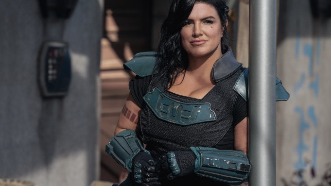 A futuristic heroine, channeling Gina Carano’s bold energy, stands outside on the grounds of an epic adventure park. With arms crossed and a confident expression, she strikes a powerful pose as if ready to lead guests on the next thrilling attraction.