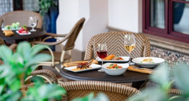 An outdoor dining table at Disneyland Resort’s Carthay Circle Lounge, set with two glasses of wine and elegantly plated dishes, surrounded by stylish woven chairs and lush greenery—evoking the sophisticated alfresco atmosphere beloved by Disney fans.