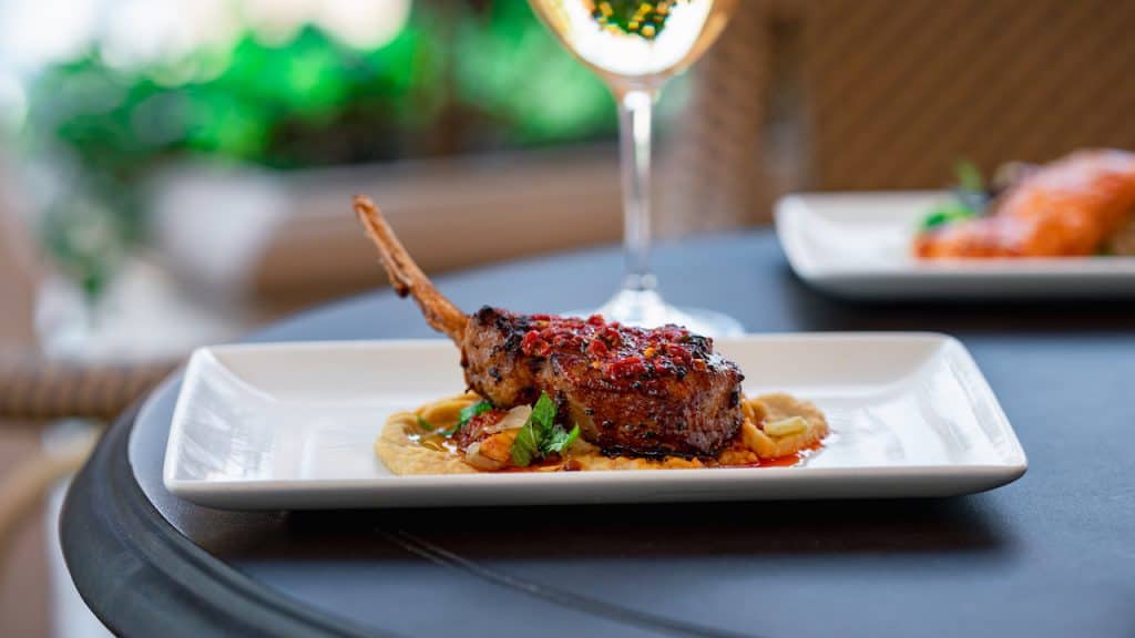 A mouthwatering grilled lamb chop rests on a creamy puree, garnished with fresh herbs and elegantly plated at Carthay Circle Lounge, with a signature Disney cocktail in the background—an ideal bite for guests savoring upscale flavors during their Disneyland adventure.