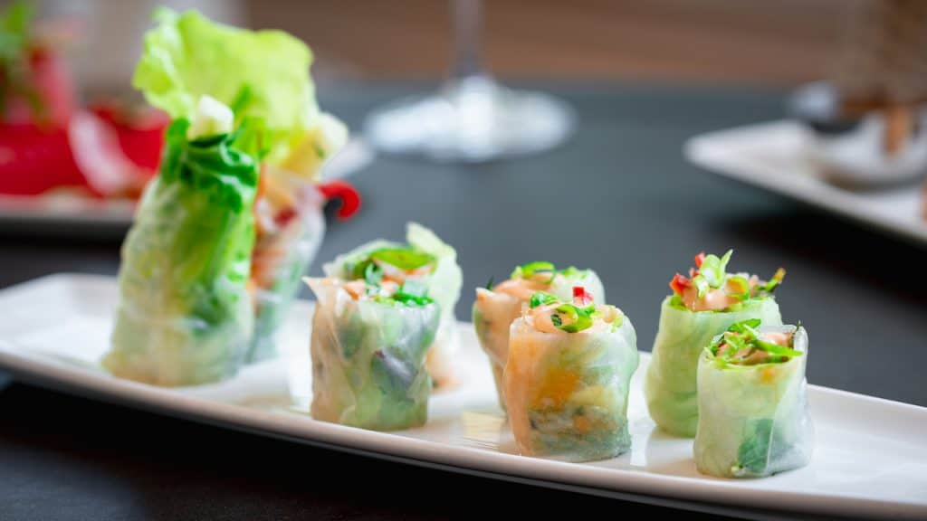 Vibrant Vietnamese spring rolls stuffed with fresh veggies are arranged on a white plate, ready to enjoy at Carthay Circle Lounge, one of Disney California Adventure’s signature dining spots. The inviting patio and softly blurred park backdrop capture the lively atmosphere that makes outdoor dining at Disneyland Resort so memorable.