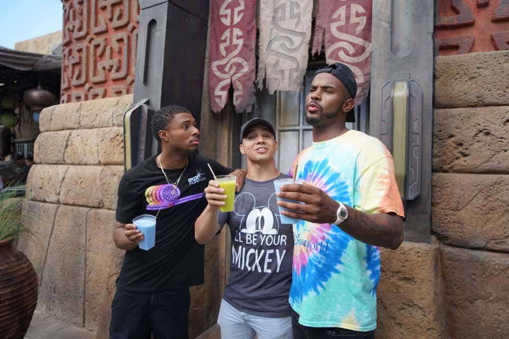 Three friends enjoy specialty drinks as they gather in front of a classic Disney Parks attraction, sharing laughter and conversation. The scene channels the chill, trendy vibe of a grown-ish episode—imagine Trevor Jackson’s energy set against the vibrant backdrop of Main Street, U.S.A., for the ultimate carefree park day.