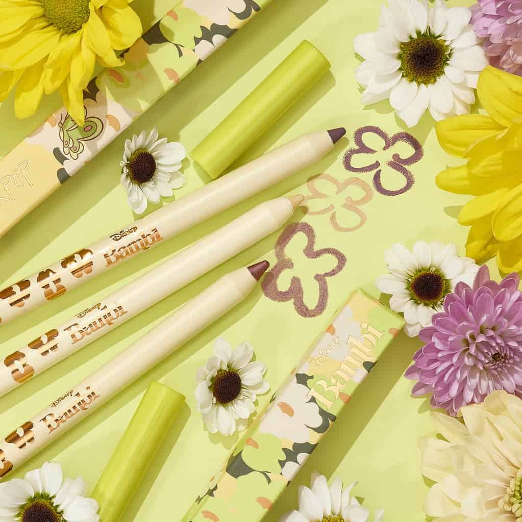 Three ColourPop Bambi-inspired makeup pencils lie against a backdrop of playful doodled blooms and real daisies and chrysanthemums, bringing the enchanting spirit of Disney’s Bambi to life with a touch of theme park whimsy.