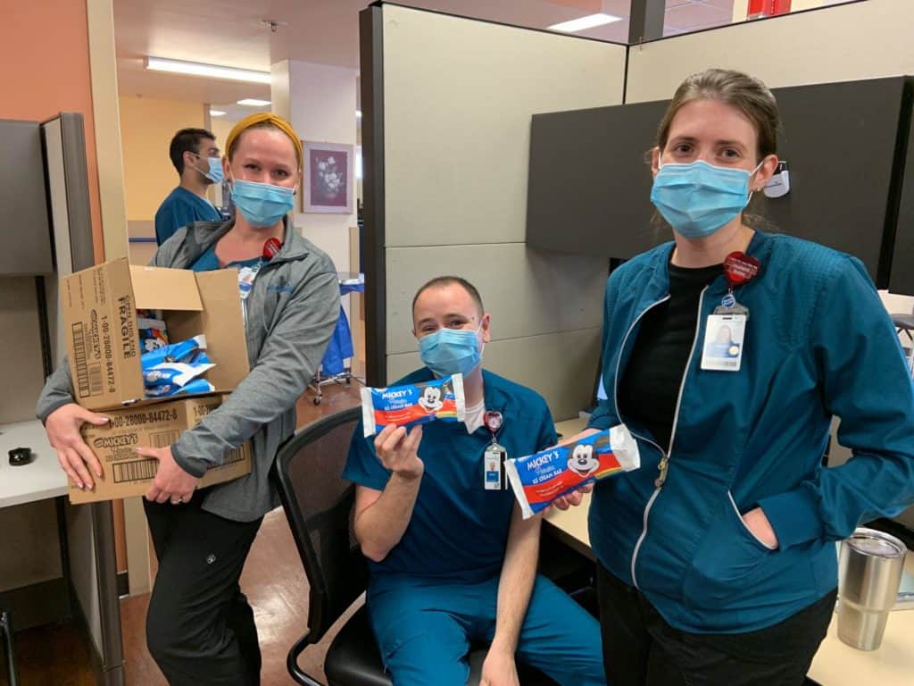 At AdventHealth Orlando, three team members dressed in scrubs and masks enjoy classic Mickey Ice Cream Bars and smile together in their office, marking the excitement of the hospital’s partnership as an official Disney Health Partner.