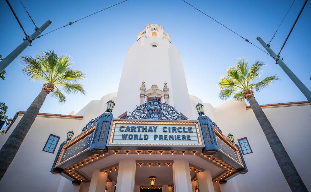 Carthay Circle Theater’s grand entrance welcomes guests at Disney California Adventure, framed by swaying palm trees and a bright blue sky. The marquee announces "Carthay Circle World Premiere," inviting visitors to enjoy alfresco dining at the Carthay Circle Lounge in a glamorous, Hollywood-inspired atmosphere.