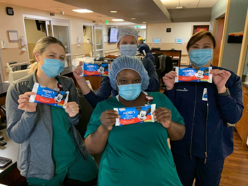 Four healthcare workers in masks enjoy iconic Mickey Mouse ice cream bars and smile for the camera inside a hospital, celebrating the magical collaboration between AdventHealth Orlando and Disney’s Health Partner.