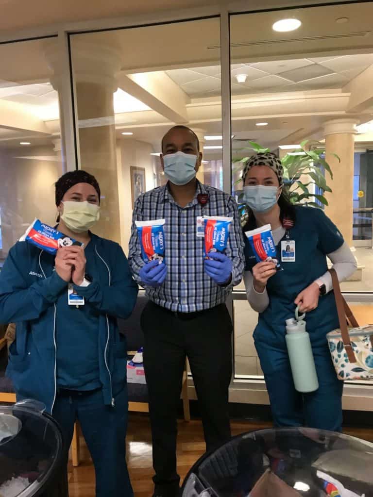 At AdventHealth Orlando, three masked team members in scrubs proudly hold up instant coffee packets as they stand together indoors, celebrating their role as part of the Disney Health Partner community.