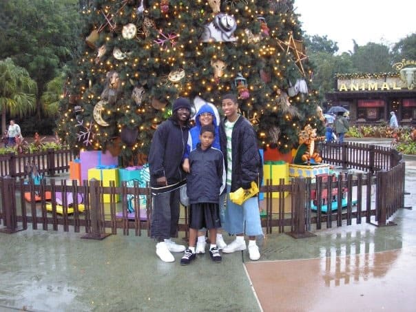 A group of friends in raincoats beams with joy in front of a grand, sparkling Christmas tree at Disney Parks, immersed in the holiday magic and festive atmosphere.