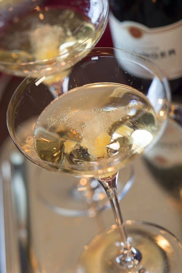 Two coupe glasses brimming with sparkling wine and garnished with lemon twists capture the sophistication of the Colette Champagne Cocktail, a signature drink at Disney Cruise Line’s Remy restaurant, glimmering on a polished surface—perfect for Disney fans seeking a touch of onboard luxury.