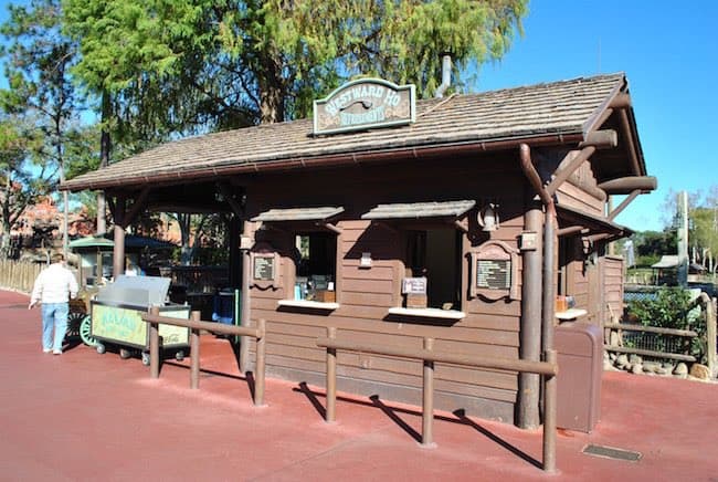 Westward Ho, the charming wooden snack stand in Magic Kingdom, sits shaded beneath leafy trees with eye-catching menu boards inviting guests to stop for a quick bite. A park-goer stands nearby, ready to order classic theme park treats.