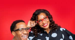 Two friends grinning in their Foster Grant reading glasses, ready for theme park adventures, with a vivid red backdrop evoking the excitement of classic amusement attractions.