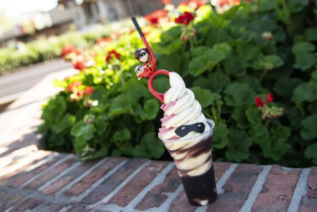 Swirled ice cream dessert featuring a black mask decoration and topped with Mrs. Incredible, served on a brick ledge surrounded by vibrant red flowers—this exclusive treat at Disney Springs is a playful nod to The Incredibles and perfect for fans seeking limited-time Pixar-inspired snacks.