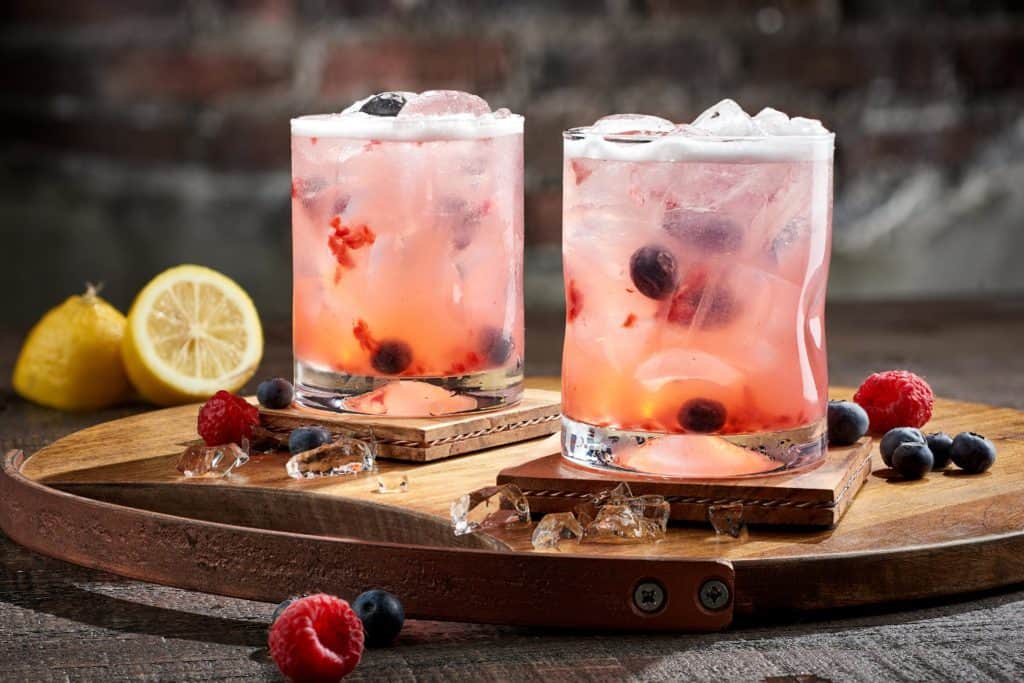 Two colorful Berry Smash Cocktails packed with ice and topped with fresh berries sit on a wooden tray, accompanied by lemon slices and extra ice—ready to be enjoyed just like you would at The Edison in Disney Springs, capturing that perfect Disney nightlife vibe.