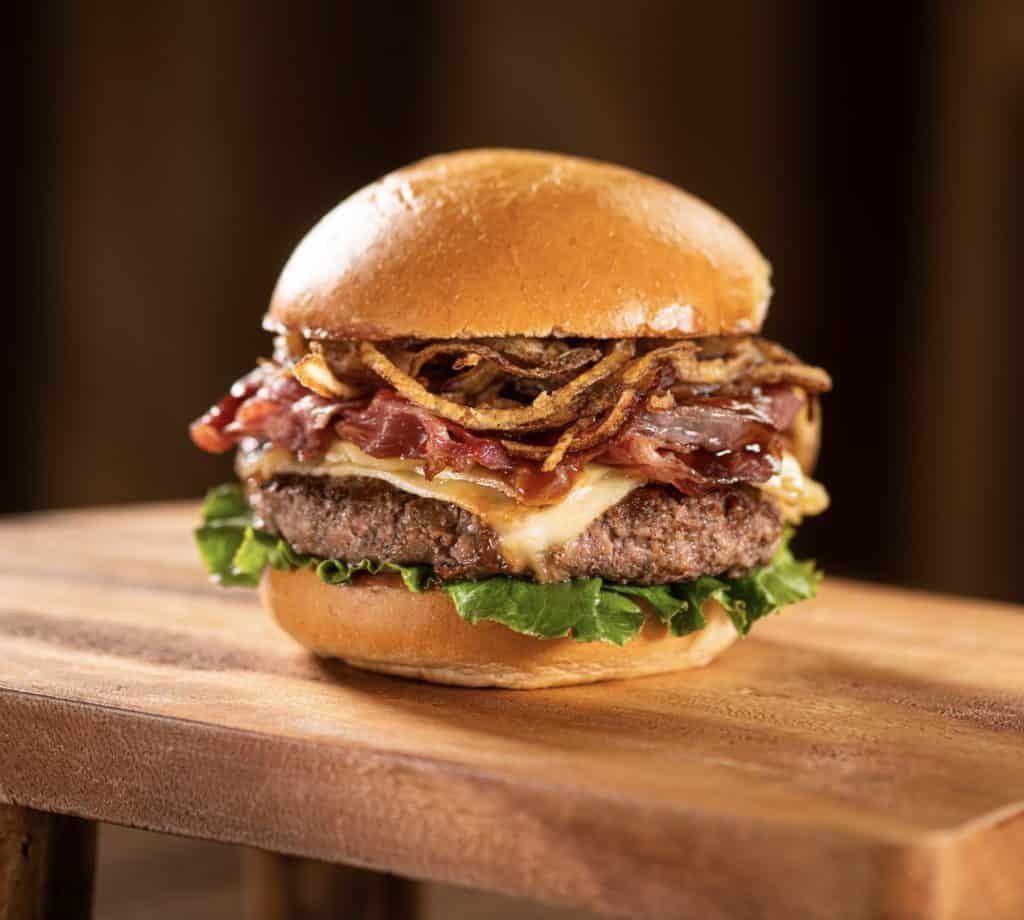 The Dubliner Burger at Disney Springs boasts lettuce, cheese, bacon, and crispy onions stacked on a freshly baked bun, all presented on a rustic wooden surface—an irresistible bite for anyone exploring the flavors of Walt Disney World’s dining scene.