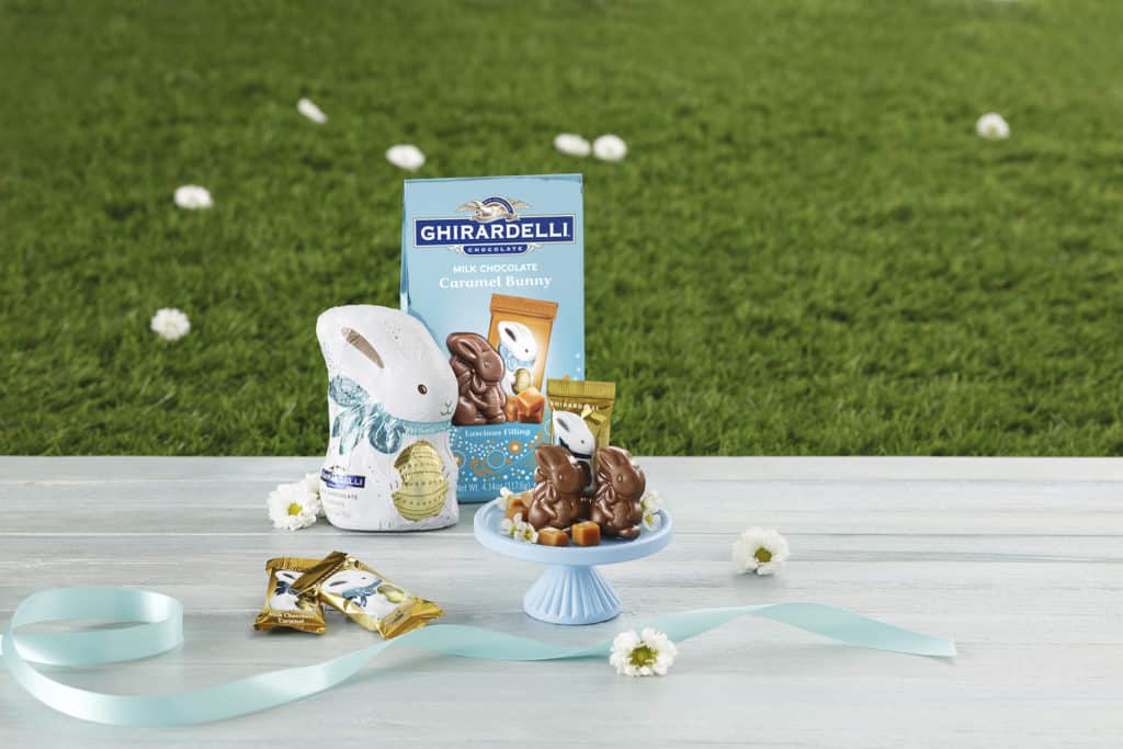 Ghirardelli caramel bunnies and chocolates, featuring cute Mini Caramel Bunnies and a classic Milk Chocolate Bunny, are artfully displayed on a blue stand adorned with ribbon and flowers against a grassy backdrop—capturing the festive spirit of Disney Springs Ghirardelli for your Easter celebration.