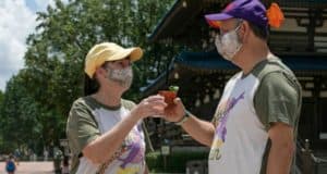 Festival-goers in coordinated shirts and masks clink their drinks in celebration outside a pagoda on a sunny day at EPCOT International Food & Wine Festival.