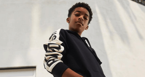 Isaiah Russell-Bailey, channeling his signature confidence from Disney+’s Crater, stands outside in a stylish black hoodie with bold white sleeve lettering, gazing directly at the camera—capturing that effortlessly cool energy you’d expect from a young Disney star.
