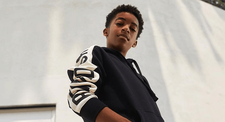 Isaiah Russell-Bailey, channeling his signature confidence from Disney+’s Crater, stands outside in a stylish black hoodie with bold white sleeve lettering, gazing directly at the camera—capturing that effortlessly cool energy you’d expect from a young Disney star.