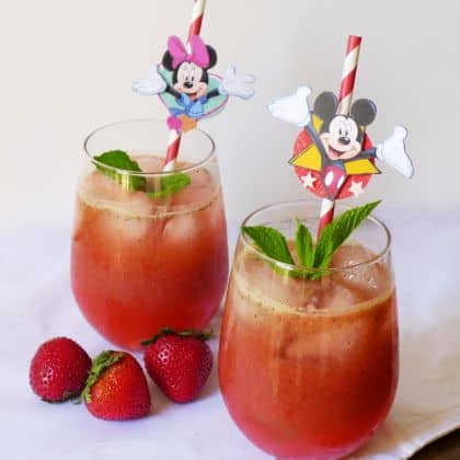 Two glasses of Strawberry Basil Mint Lemonade with ice, fresh mint, and fun character straws sit beside vibrant strawberries on a festive table—an ideal treat to enjoy while taking a break at your favorite theme park.