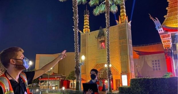 Guests in masks chat beneath swaying palm trees, set against the dazzling nighttime backdrop of the illuminated Chinese Theatre and its iconic spires at Disney’s Hollywood Studios, highlighting the park’s spectacular evening lighting.