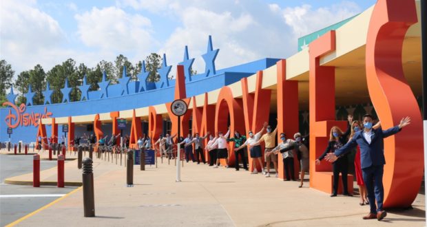 Guests wearing masks are happily welcomed back to Disney’s All-Star Movies Resort, posing in front of the iconic sign as Disney Cast Members greet everyone with cheerful waves and safety measures under the sunny sky.