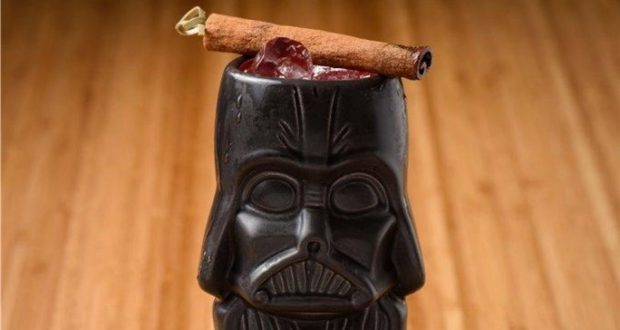 Darth Vader mug in all its galactic glory, filled with a dark iced Darth 'N Stormy cocktail and topped with a cinnamon stick lightsaber, ready to tempt you over to the dark side at Star Wars: Galaxy’s Edge, served atop a rustic wooden table.