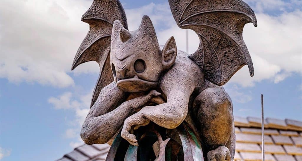 A winged stone gargoyle perches atop a rooftop beneath a brilliant, cloud-dappled sky, evoking the darkly enchanting atmosphere that fans adore at Gideon's Bakehouse in Disney Springs.