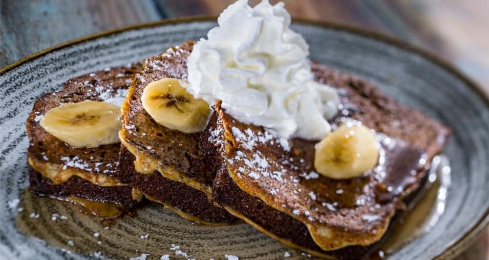 Indulge in three slices of French toast piled high with fresh banana slices, a dusting of powdered sugar, and fluffy whipped cream—an irresistible Easter Brunch treat you can enjoy at Walt Disney World restaurants.