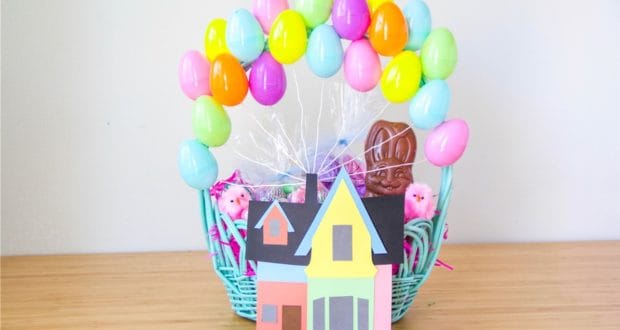 Blue Up Easter basket display styled like a whimsical theme park attraction, featuring balloon-like colorful eggs, a mini paper house reminiscent of parade floats, a chocolate bunny as the star character, and vibrant Easter grass set atop a wooden boardwalk.