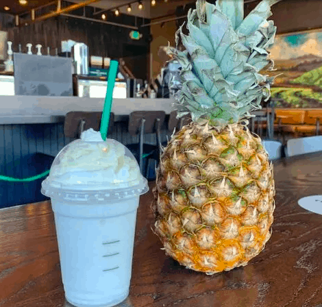 A Starbucks Dole Whip Frappuccino topped with whipped cream sits beside a fresh pineapple on a wooden table inside a coffee shop, evoking the fun and tropical flavors beloved by Disney park fans.