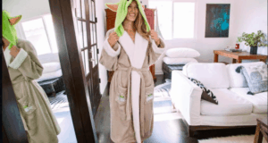 Grogu, dressed in a cozy bathrobe, beams with a joyful smile inside a sleek, light-filled lounge, his reflection visible in a nearby mirror.