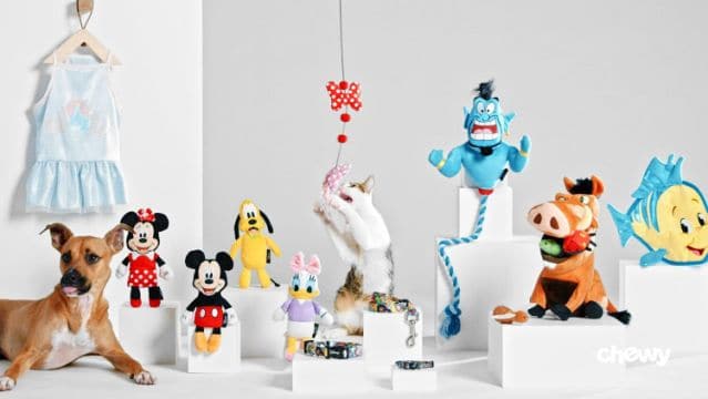 Dog and cat enjoying Disney magic with plush toys from the Chewy Disney Collection, a princess-style blue dress on display, and stylish collars arranged like character accessories; Chewy logo featured—making every pet feel like they're part of the happiest place on earth.