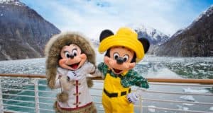 Mickey and Minnie Mouse, dressed in festive winter attire, greet guests on the deck of a Disney Cruise Line ship surrounded by sparkling icy mountains and serene waters, highlighting the thrilling new summer 2022 destinations for Disney fans.