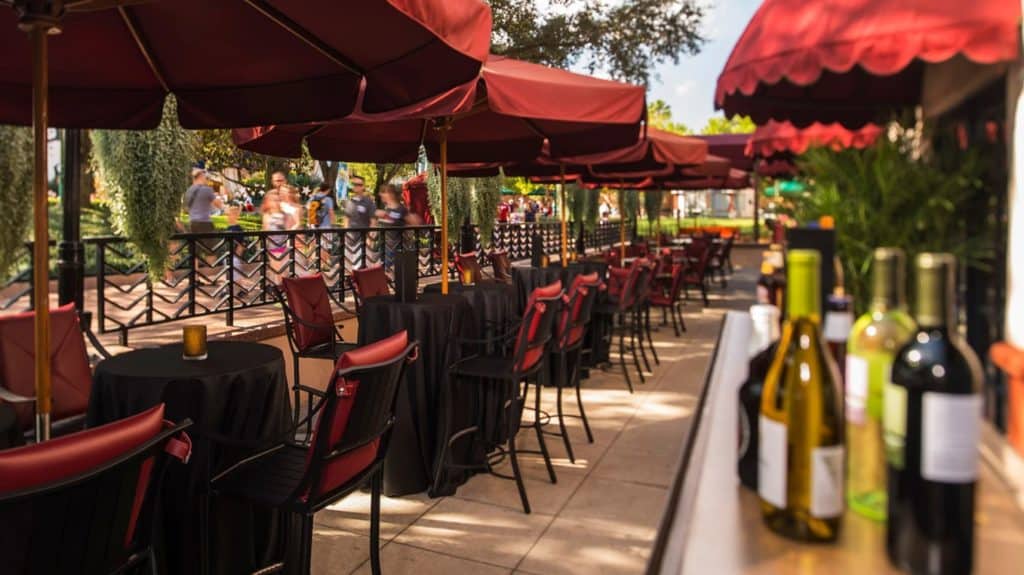 An outdoor dining patio at Disney World, featuring vibrant red umbrellas shading tables and chairs, with bottles of wine set up in the foreground—capturing the magic of Disney’s lively table-service restaurant atmosphere, perfect for guests enjoying al fresco meals in true theme park style.