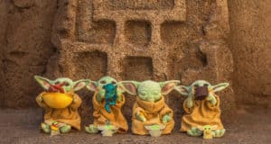Grogu figures dressed in classic tan robes are displayed against a textured backdrop, each holding colorful plushes and collectible pins—perfect photo ops for Star Wars fans at Galaxy’s Edge.