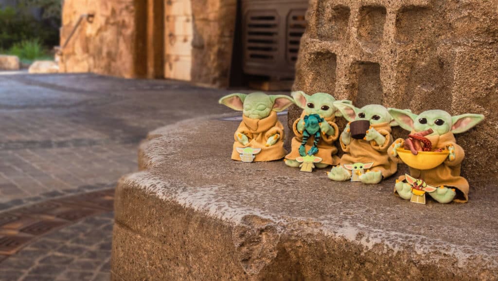 Grogu plushes line up on a stone ledge at Star Wars: Galaxy’s Edge, each holding unique souvenirs with Black Spire Outpost’s iconic buildings in the background—the ultimate collectible set for Mandalorian fans, especially when matched with limited-edition Grogu pins from the park.