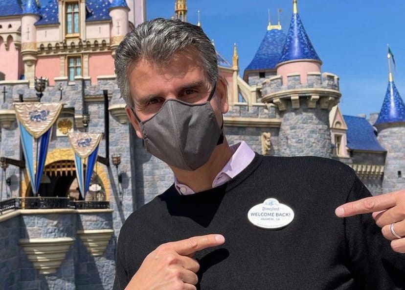 A Disneyland guest wearing a mask enthusiastically points to a “Welcome Back!” badge in front of Sleeping Beauty Castle, capturing the excitement of the park’s highly anticipated reopening.