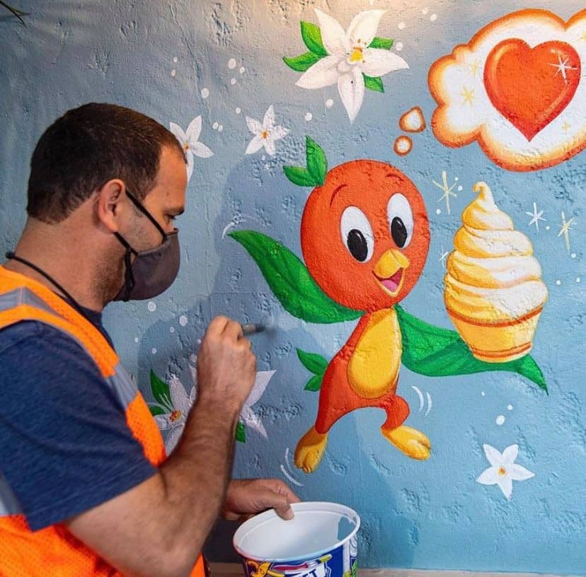 An artist brings Magic Kingdom magic to life by painting a bright new mural starring Orange Bird, enjoying an ice cream and surrounded by playful flowers and a heart-shaped thought bubble.