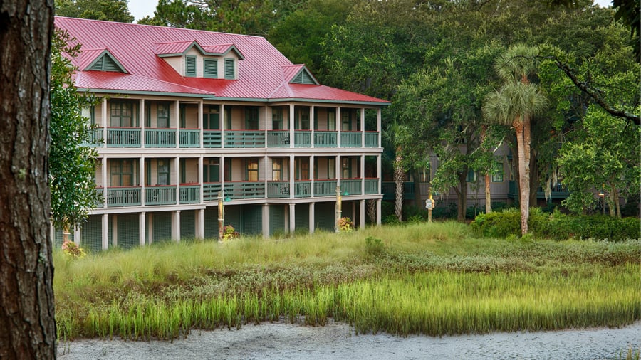 A three-story building with a red roof and green balconies stands surrounded by lush trees and tall grass near the water at Disney’s Hilton Head Island Resort—a scenic retreat where Disney fans can celebrate milestone anniversaries and enjoy resort-style relaxation in a picturesque, nature-filled setting.