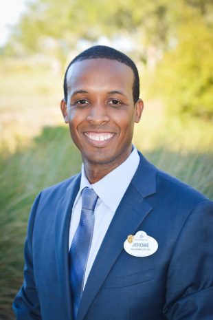 A smiling Disney cast member in a sharp blue suit and tie, name tag proudly displayed, greets guests outside amid lush greenery at Disney’s Hilton Head Island Resort.