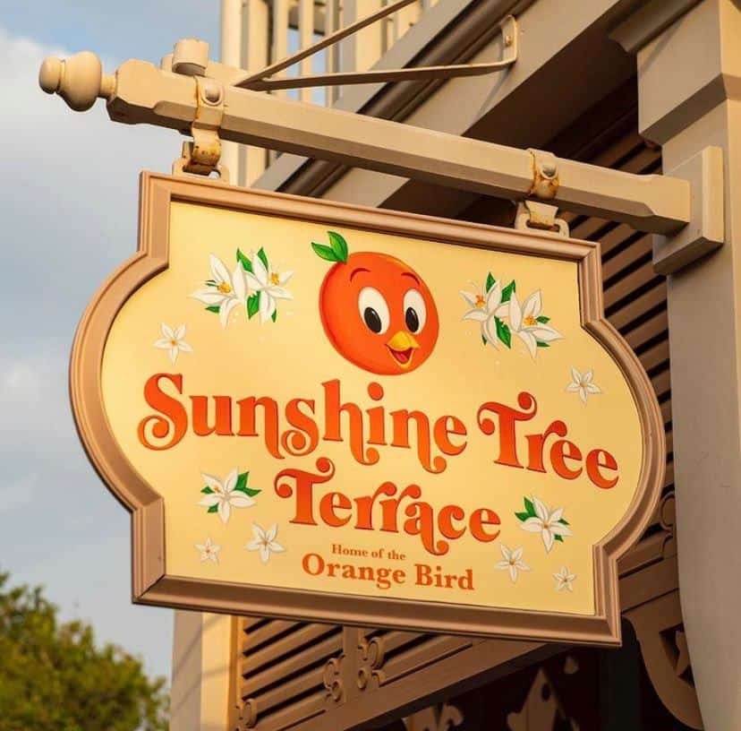 The Sunshine Tree Terrace sign, featuring the beloved Orange Bird and accented with white flowers, hangs outside its location in Magic Kingdom, just steps from the park’s vibrant new mural.