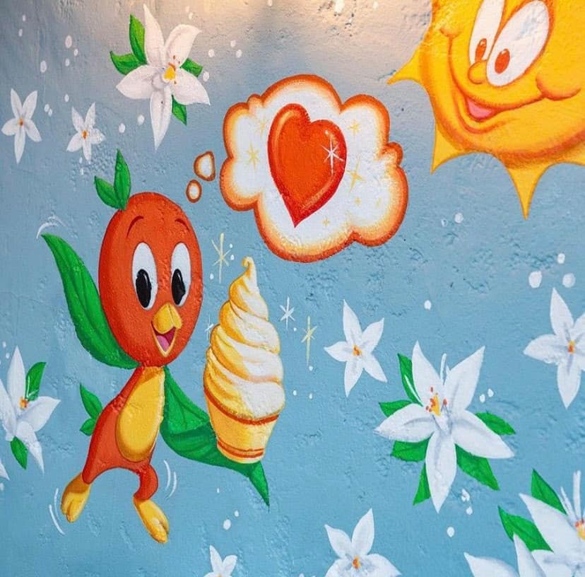Orange Bird cheerfully holds a swirl of soft serve ice cream, set against a vibrant blue mural bursting with colorful flowers and a beaming sun—this upbeat new addition channels classic Magic Kingdom nostalgia, instantly transporting guests to the heart of Walt Disney World magic.