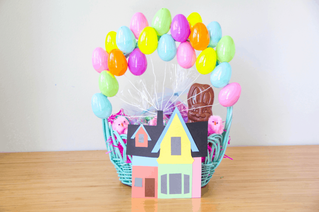 A blue basket filled with colorful balloons, a chocolate bunny, and a paper house sits on a wooden table—an Up-themed Easter basket designed to capture the wonder of Pixar's Up experience at Disney Parks.