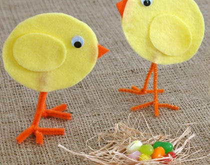 Two playful yellow felt chicks with vibrant orange legs, evoking the charm of Cinderella's Easter celebration at a beloved theme park, gaze at a nest brimming with colorful jelly beans on rustic burlap—a whimsical touch perfect for bringing Disney-inspired Easter magic to any table display.