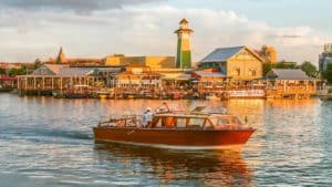 A classic wooden boat glides across the lake at Walt Disney World, passing colorful waterfront buildings and a picturesque lighthouse—capturing the magical atmosphere of Disney’s iconic table-service restaurants with their welcoming outdoor patios.