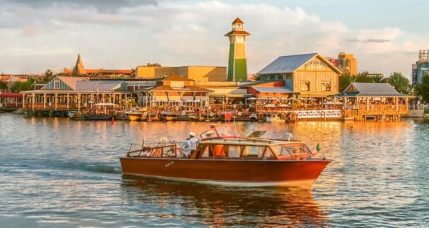 A classic wooden boat glides across the lake at Walt Disney World, passing colorful waterfront buildings and a picturesque lighthouse—capturing the magical atmosphere of Disney’s iconic table-service restaurants with their welcoming outdoor patios.