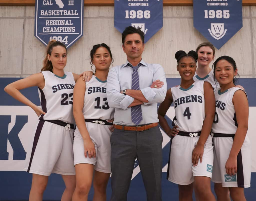 A basketball coach and five players, reminiscent of the spirited team from Disney+’s Big Shot, pose together in front of championship banners inside a gym—channeling the determination and camaraderie celebrated in Disney sports stories.