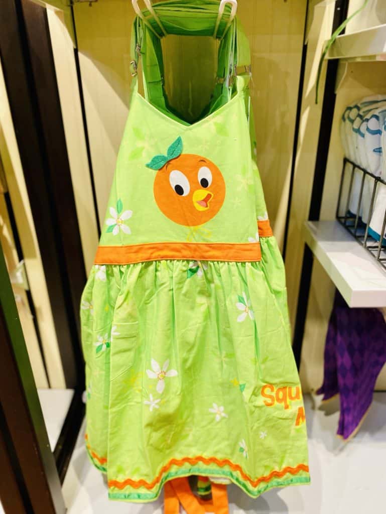 Green apron featuring an orange cartoon face with leafy accents, white flowers, and orange trim, displayed on a rack—ideal for Disney Parks fans who love whimsical merchandise inspired by iconic characters.