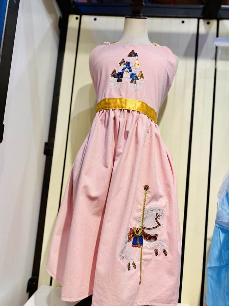 Pink dress featuring enchanting embroidered castles and carousel horses, capturing the whimsical charm of Disney Park fashion, showcased on a mannequin.